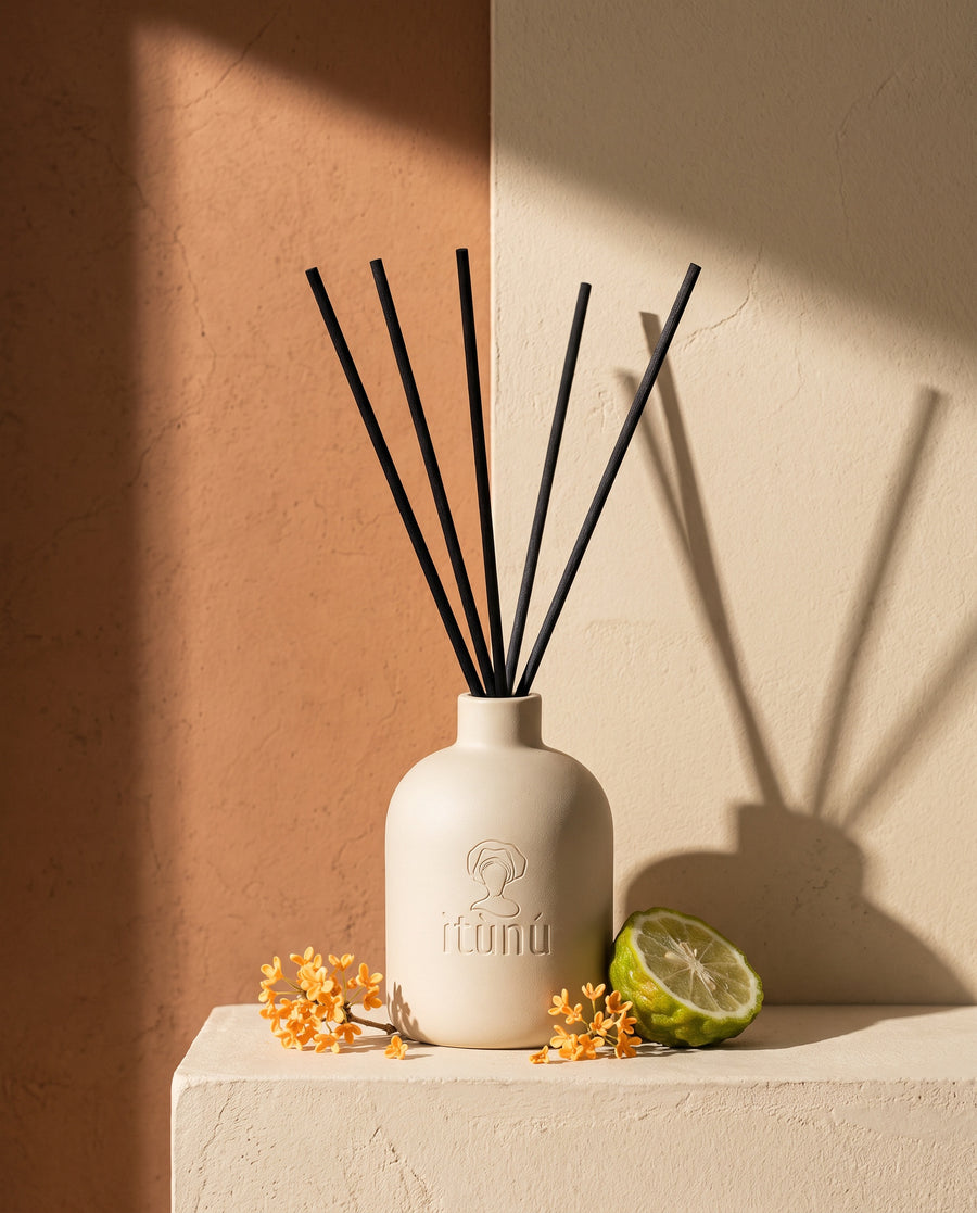The Ìtùrá reed diffuser on a wheat plaster plinth against a terracotta and wheat wall, with osmanthus flowers and half a bergamot
