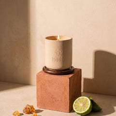The lit Ìtùrá candle on its walnut coaster, standing on a terracotta cube against a wheat wall, with a cut bergamot and pieces of amber resin at its base