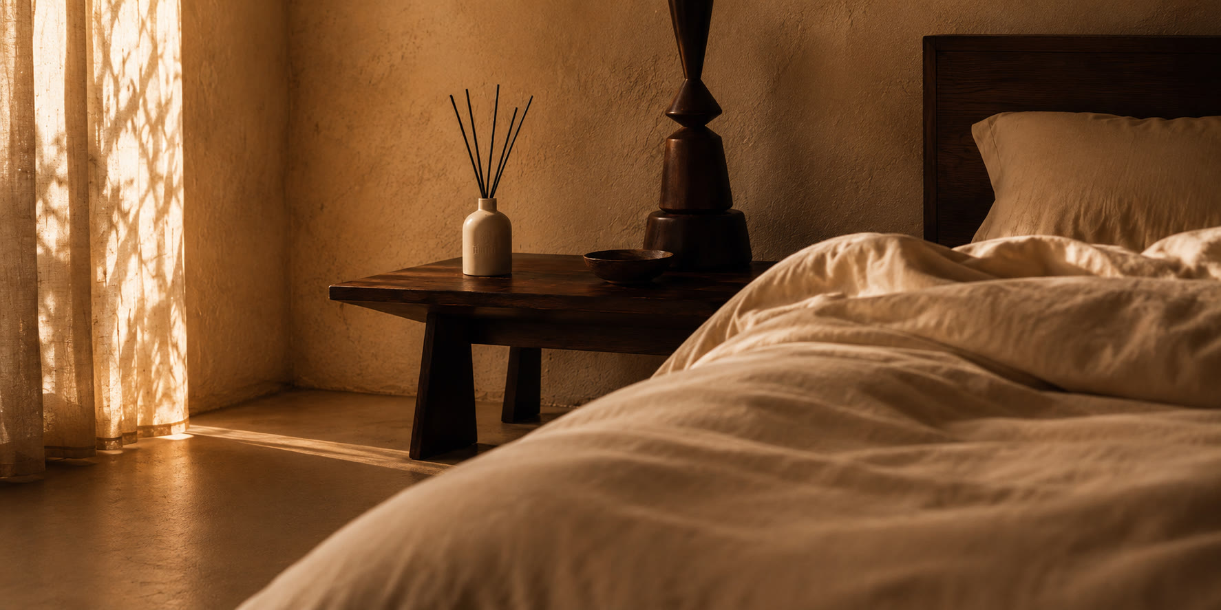 A bedroom corner at the end of the day. A cream reed diffuser with dark reeds on a dark wood nightstand beside an unmade bed in cream linen, low light through a curtain.