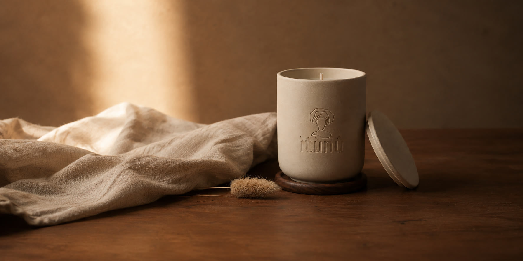 An unlit cream ceramic ìtùnú candle, its lid resting against the base, on a round walnut coaster on a dark wood table, rumpled linen gathered to the left and a dried grass stem lying in front.