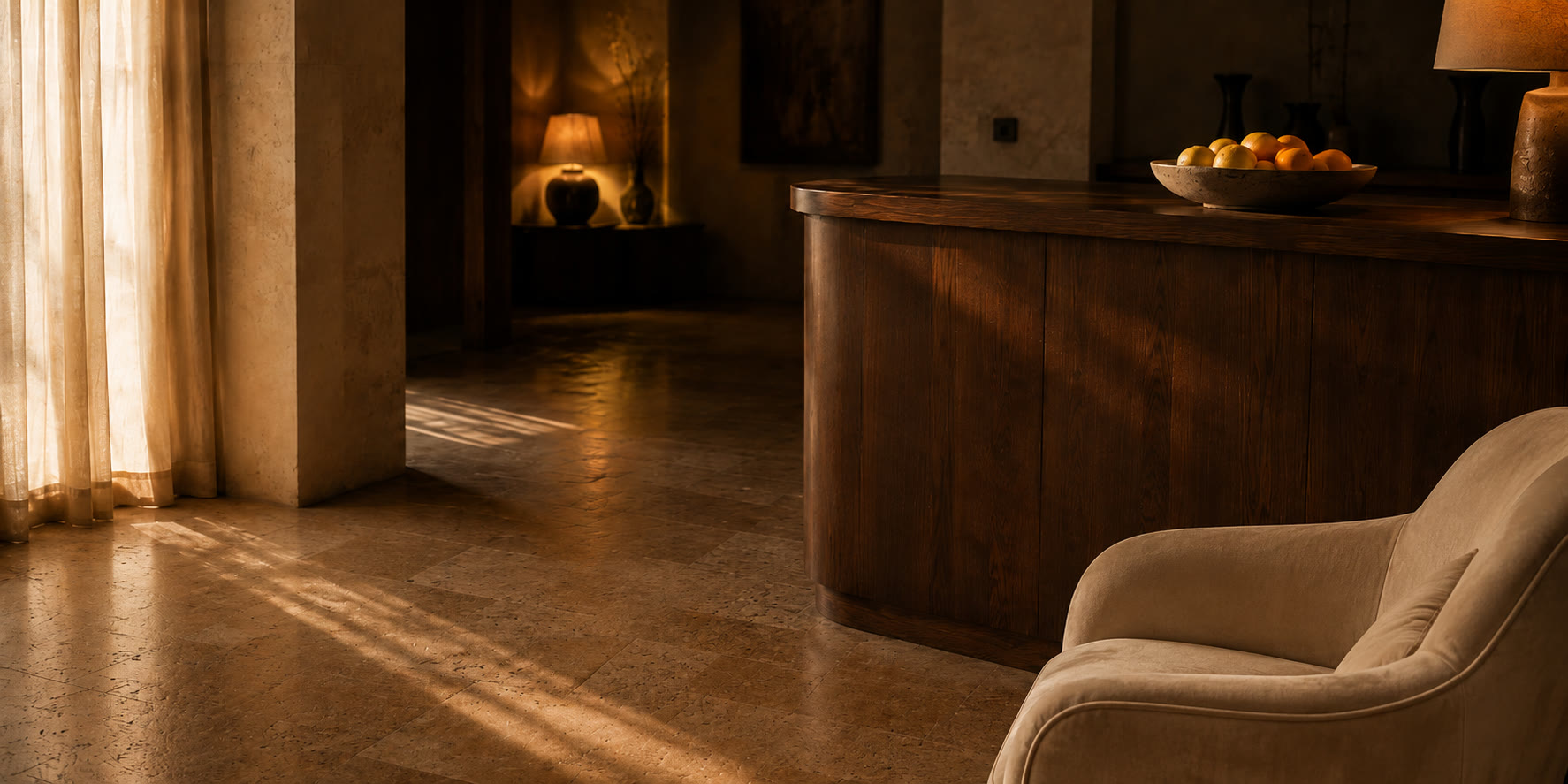A hotel entrance hall at dusk. A shallow stone bowl of oranges sits on a curved walnut concierge desk, honey travertine floor, light falling through a sheer curtain at the left.