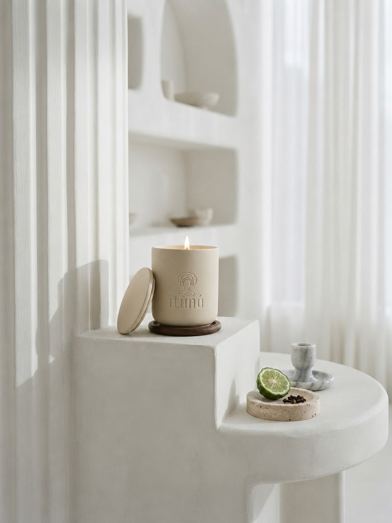 A lit Ìtùnú candle on a walnut coaster, its lid leaning against it, on a white plaster step beside sheer curtains, with a halved bergamot on a small stone dish below.