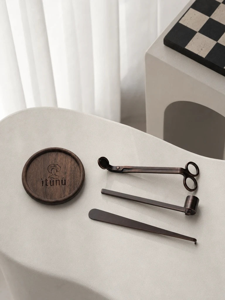 The walnut Ọ̀nà coaster and the bronze wick trimmer, snuffer and aligner on a white cloud-shaped table, a chess board on a plaster console behind