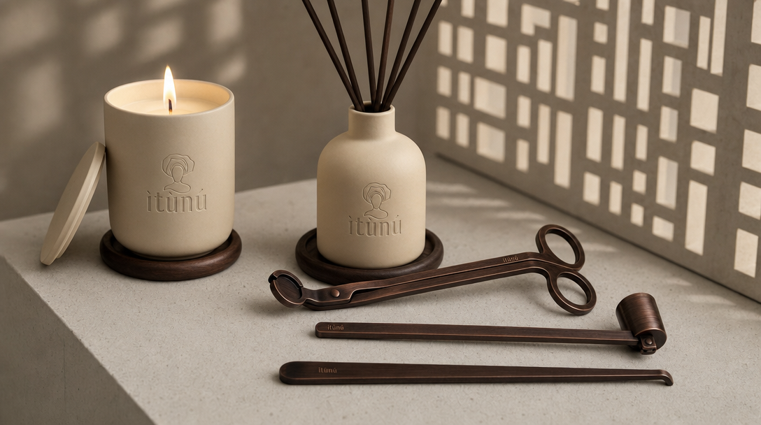 The complete set on a pale stone ledge: the lit cream ceramic candle with its lid leaning against the base on a walnut coaster, the matching reed diffuser with black reeds on a second coaster, and the three bronze Ọ̀nà tools laid in front. A perforated screen throws a grid of light on the wall behind.