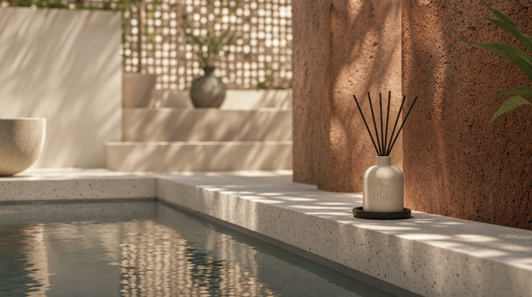 The diffuser small on a terrazzo ledge beside a still reflecting pool in a courtyard of ochre laterite piers and pale screen walls, dappled light across the stone.