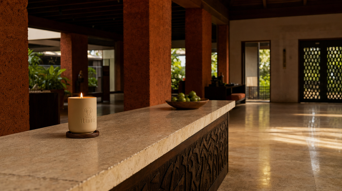 The lit Ìtùrá candle small on a long stone reception desk in a hotel lobby of ochre laterite piers and carved timber, a shallow bowl of green bergamots further along and polished travertine stretching away.