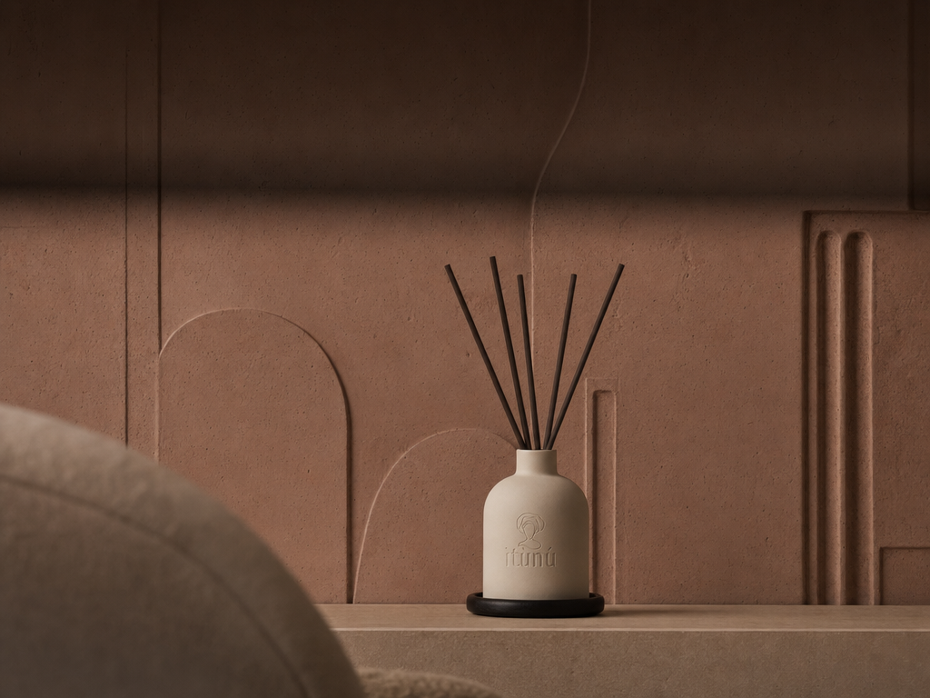 The diffuser on a low ledge against a terracotta wall carved with soft arched reliefs, the shoulder of an upholstered armchair soft in the foreground.