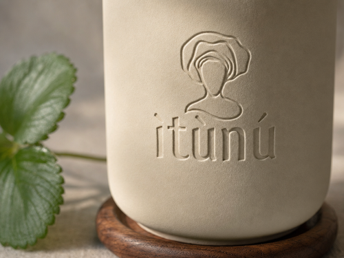 Macro of the debossed ìtùnú mark and wordmark on the matte cream ceramic, raking light picking out the relief, a green strawberry leaf soft at the left edge and the walnut coaster below.