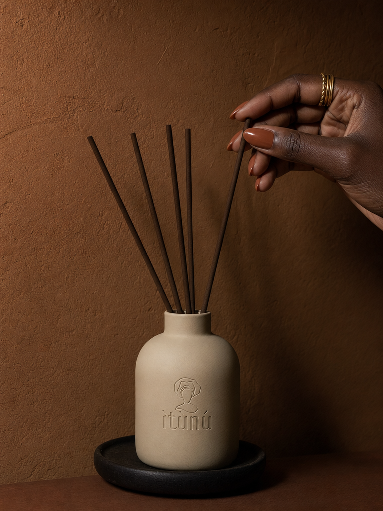 A hand with gold rings lifts one black reed from the cream ceramic diffuser, which stands on a black dish against a deep terracotta wall.