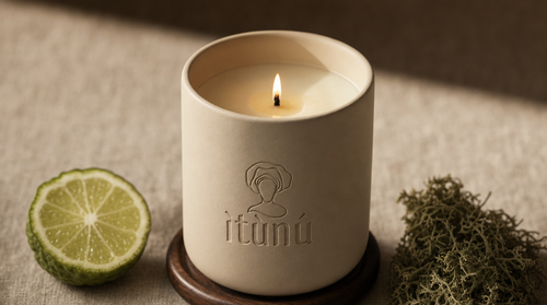 Close view of the lit Ìtùrá candle on its walnut coaster, a halved bergamot on the left and a knot of pale oak moss on the right, on natural linen.