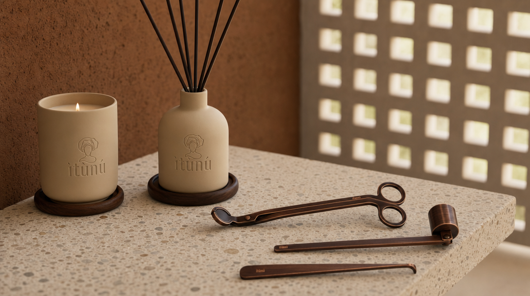 The complete set on a terrazzo counter against an ochre laterite wall: the lit cream ceramic candle on a walnut coaster, the matching reed diffuser with black reeds on a second coaster, and the three bronze Ọ̀nà tools laid in front. A perforated screen wall stands behind in bright daylight.