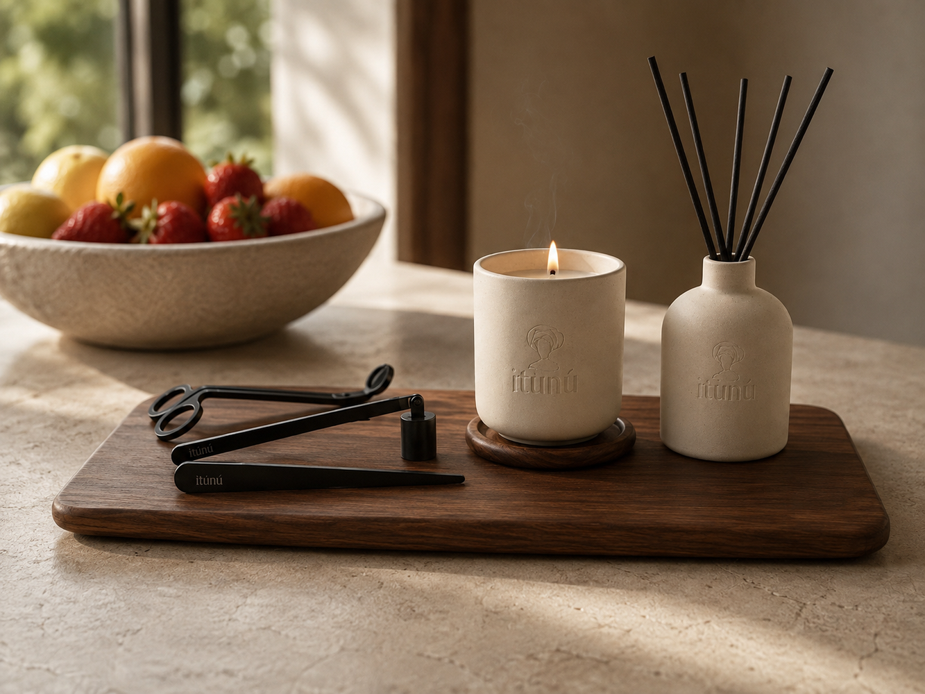 The Ìtọ́jú set on a walnut tray: a lit cream ceramic candle on a round walnut coaster, the matching reed diffuser with black reeds, and the three black Ọ̀nà tools: the scissor wick trimmer, the snuffer with its cylindrical cup, and the tapered wick aligner. A bowl of lemons, oranges and strawberries sits behind on a travertine counter.