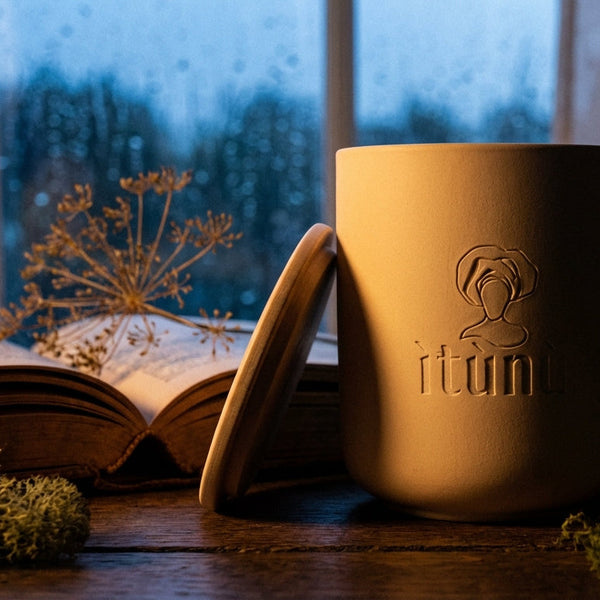 Matte cream ceramic candle with ìtùnú branding on a wooden surface near an open book and plants, with a window in the background.