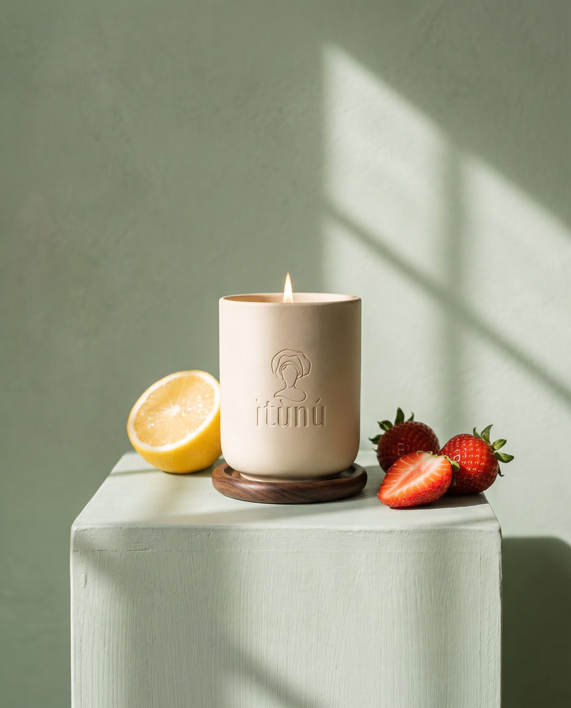 The lit Ìtọ́jú candle on its walnut coaster on a celadon block against a sage wall, with half a lemon and fresh strawberries in dappled light