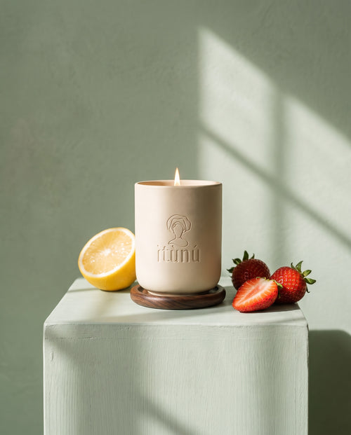 The lit Ìtọ́jú candle on its walnut coaster on a celadon block against a sage wall, with half a lemon and fresh strawberries in dappled light