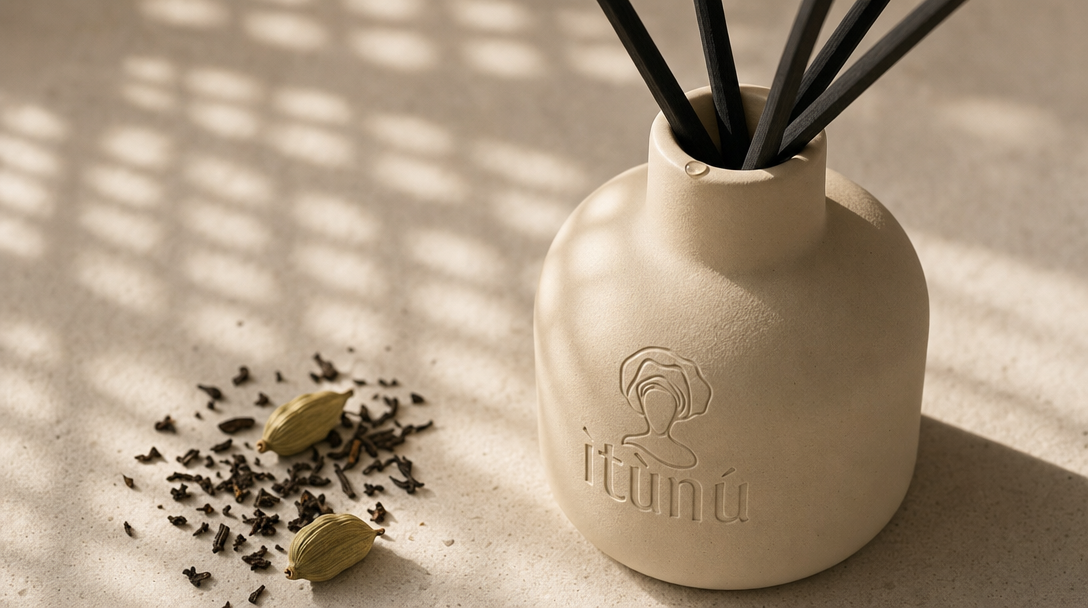 Close view of the cream ceramic diffuser's shoulder and neck with black reeds, a scatter of crushed dark tea leaves and two green cardamom pods on the pale stone beside it, grid light falling across the surface.