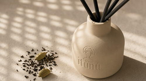 Close view of the cream ceramic diffuser's shoulder and neck with black reeds, a scatter of crushed dark tea leaves and two green cardamom pods on the pale stone beside it, grid light falling across the surface.