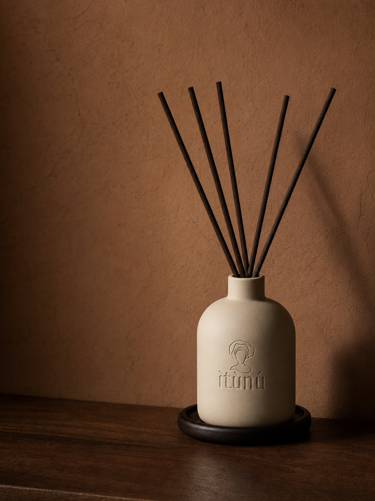 The Ìtùrá reed diffuser, a matte cream ceramic bottle with the debossed ìtùnú mark, holding five straight black reeds, standing on a dark walnut coaster on a wooden ledge against a warm terracotta wall.