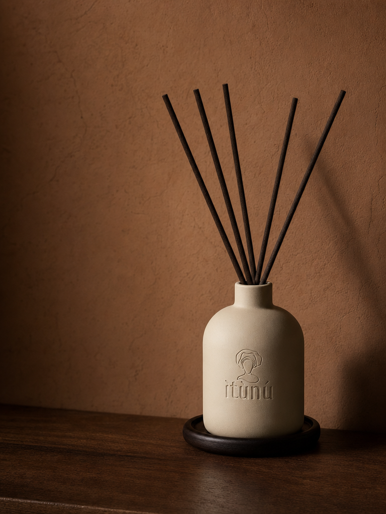 The Ìtùrá reed diffuser, a matte cream ceramic bottle with the debossed ìtùnú mark, holding five straight black reeds, standing on a dark walnut coaster on a wooden ledge against a warm terracotta wall.
