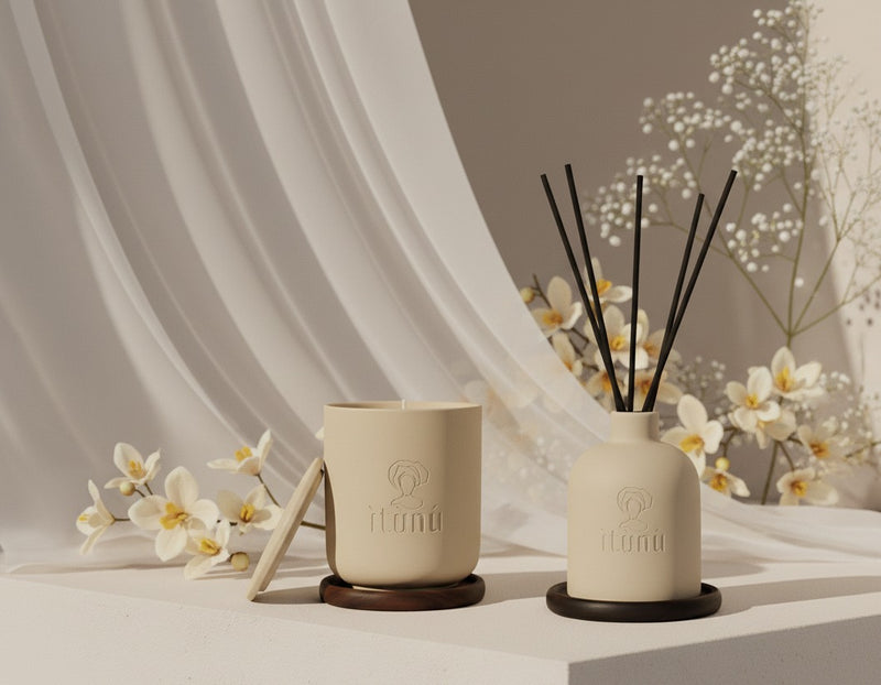 An Ìtùnú candle and an Ìtùnú reed diffuser, each on a walnut coaster, side by side on a pale plinth with small white flowers and a sheer drape behind.