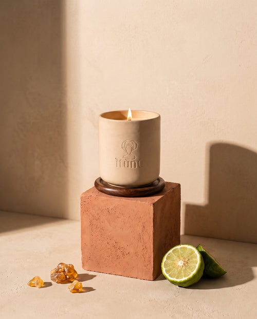 The lit Ìtùrá candle on its walnut coaster, standing on a terracotta cube against a wheat wall, with a cut bergamot and pieces of amber resin at its base