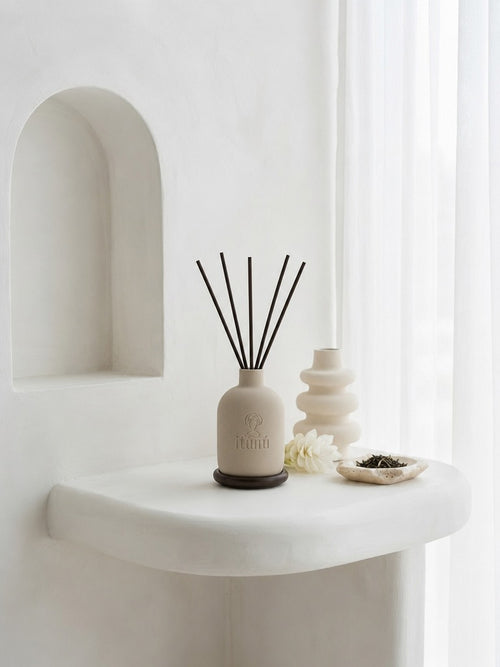 An Ìtùnú reed diffuser with black reeds on a walnut coaster, on a curved white plaster shelf by sheer curtains, with a stacked ceramic vase and a small dish of loose tea.