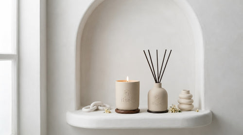 A lit Ìtùnú candle on a walnut coaster and an Ìtùnú reed diffuser with black reeds, side by side in a white plaster arched niche, with a ceramic chain link and a stacked ceramic vase beside them.