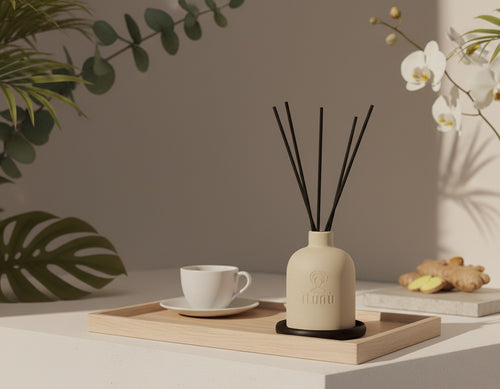 An Ìtùnú reed diffuser on a walnut coaster on a light wood tray beside a white teacup, with fresh ginger, white orchids and palm leaves around it.