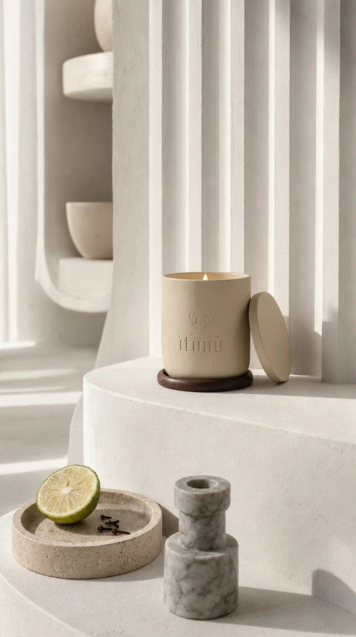 The lit Ìtùrá candle on its walnut coaster beside a fluted white plaster wall, a halved bergamot and cloves on a travertine dish and a marble candlestick below