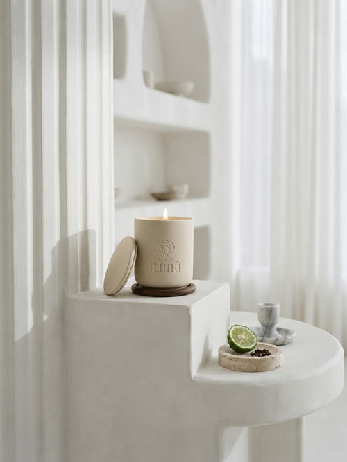 A lit Ìtùnú candle on a walnut coaster, its lid leaning against it, on a white plaster step beside sheer curtains, with a halved bergamot on a small stone dish below.