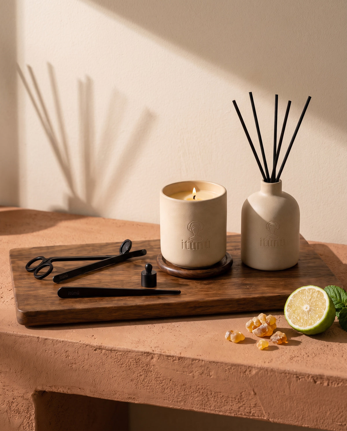 The Ìtùrá Complete Set on its walnut tray, the candle, diffuser and black tools on a terracotta bench against a wheat wall, with bergamot and amber resin